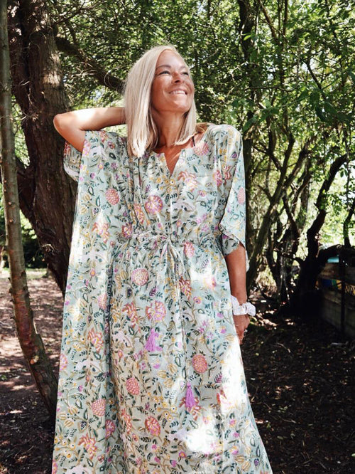 Woman standing outdoors among trees wearing Eden bohemian floral-print green cotton maxi kaftan//dress free size with relaxed sleeves and a cinched waist, smiling and looking upward.